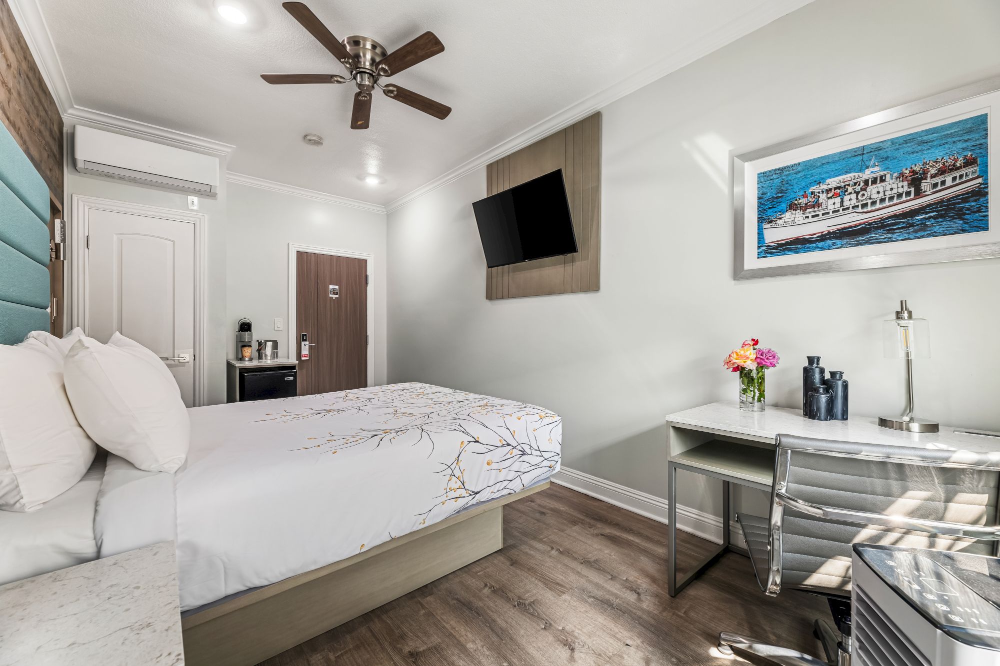 Modern hotel room: bed with white linens, wall-mounted TV, ceiling fan, desk and chair, framed ocean photo, flowers, and a small sitting area.