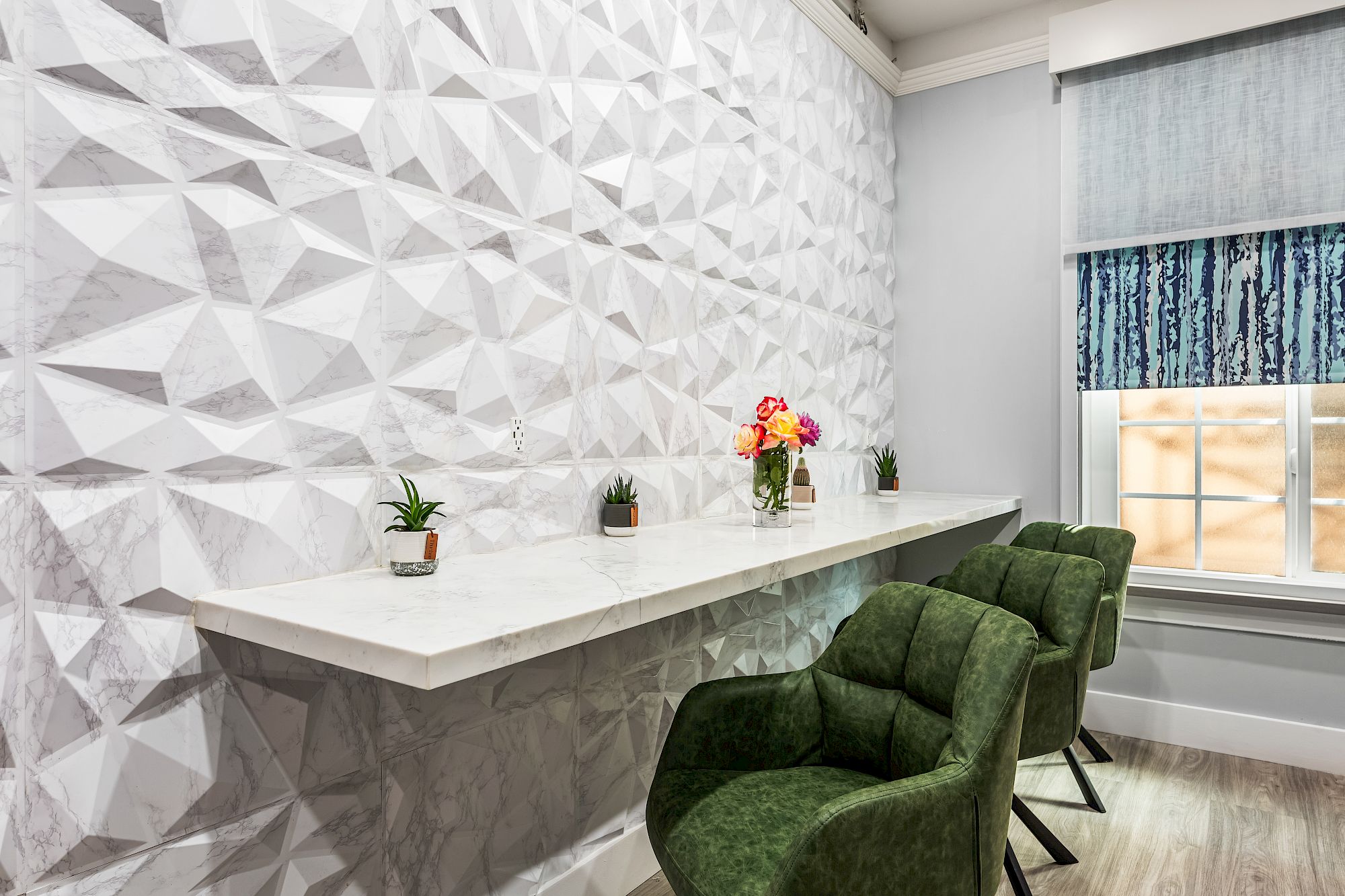 A modern lounge with a geometric 3D wall, a long white counter, green upholstered chairs, small potted plants, and a window with a patterned blind.