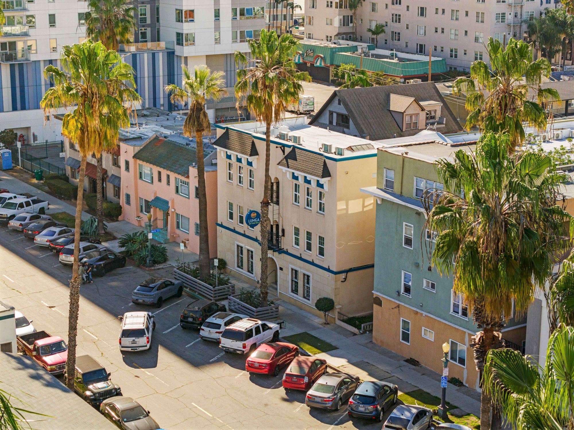 Colorful beachfront apartments with palm trees line a street, parked cars fill the lot, and sunny vibes shine over the neighborhood.