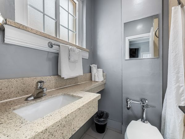 A small, modern gray bathroom with a speckled beige countertop, single sink, wall mirror, towel rack, window, and a compact toilet area.