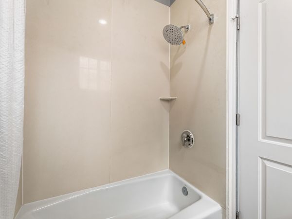 A small bathroom shower area with a white bathtub, a round showerhead, a corner shelf, beige walls, and a white door next to the tub.
