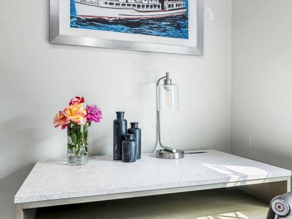 A bright, minimal home office with a marble desk, a vase of colorful flowers, black accessories, and a framed boat painting on the wall, illuminated by soft natural light.