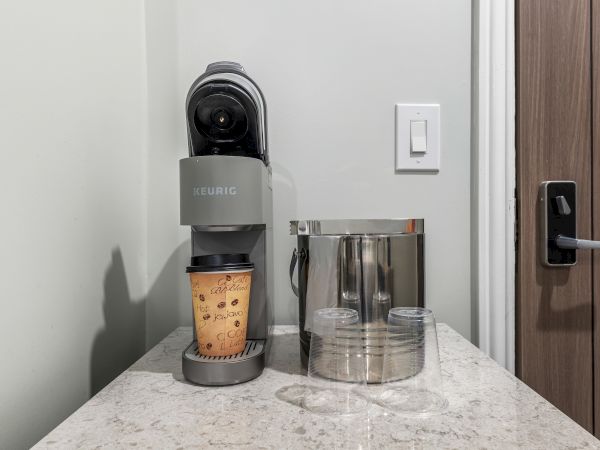 A compact espresso machine with a cup of coffee brewing, plus a metal pitcher and two clear hollow cups on a marble counter near a doorway.