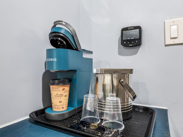 A compact coffee station with a blue espresso machine, a metal pitcher, stacked cups, a small digital control panel, and a spill tray.