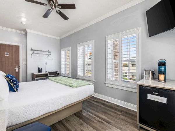 A bright hotel-style bedroom with a king bed, ceiling fan, gray walls, multiple windows with shutters, a wall-mounted TV, and a small minibar area to the right.