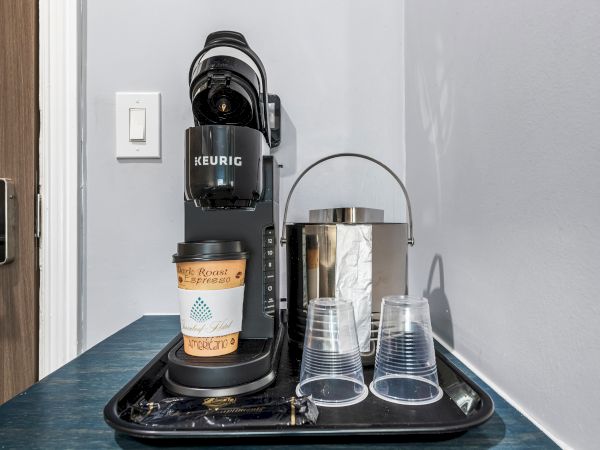 A Keurig coffee maker with a cup, a metal carafe, two plastic cups, and a small tray on a blue counter, near a light switch by a door.
