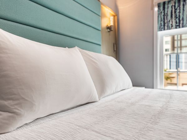 A neatly made hotel bed with white linens, soft pillows, and a teal padded headboard, near a window with city light spilling in.