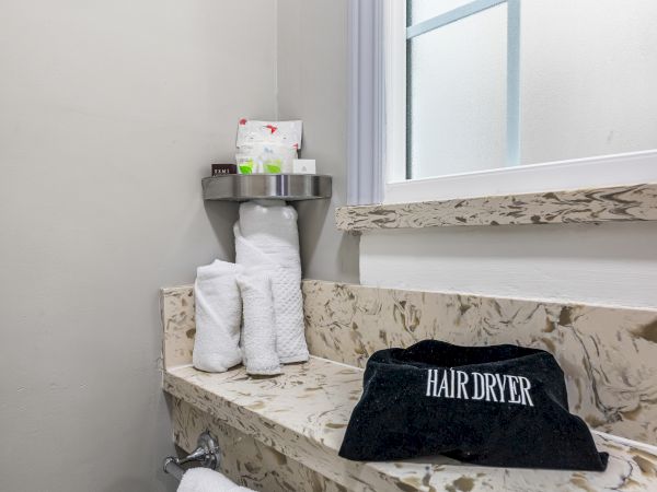 A marble bathroom counter with rolled towels, a small shelf of toiletries, a window, and a black "HAIR DRYER" case on the counter.