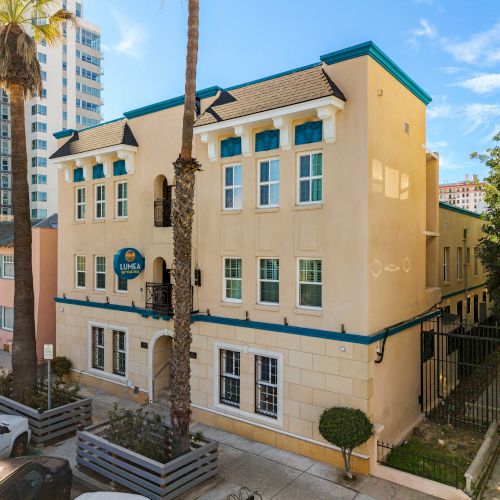 A yellow pastel building with blue trim, tall windows, palm trees, and a surrounding urban block under a bright blue sky.
