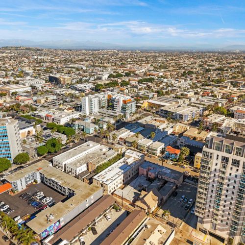 Aerial city view of a dense urban landscape with mid/high-rise buildings, scattered streets, and a sunny blue sky over a sprawling downtown.