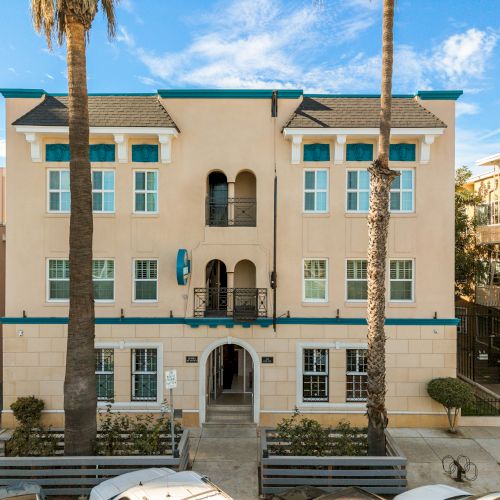 A peach-colored, coastal-style building with arched entrance, tall palm trees, and a few balconies, nestled between other pastel homes.