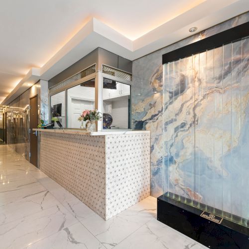 A modern hotel lobby with a long front desk, marble floors, soft lighting, and a large water feature or mural on the right wall.