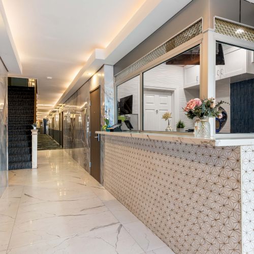 A modern hotel lobby or reception area with a long marble floor, patterned front desk, mirrored walls, soft lighting, and fresh flowers.