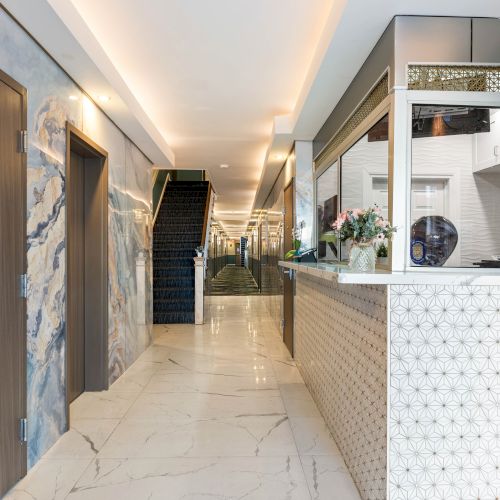 A bright hotel lobby with a marble floor, blue patterned walls, a reception desk on the right, doors along the left, and a stairway at the end.