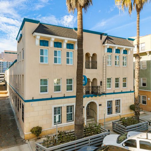 A three-story beige coastal building with arched entrance, blue trim, palm trees, and parked cars in front, sunny sky overhead.