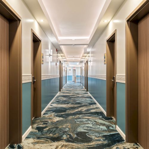 A hotel hallway with closed doors on both sides, teal wainscoting, ornate carpet, and numbered plaques along the walls. It&rsquo;s long and well-lit, ending with a bright doorway.