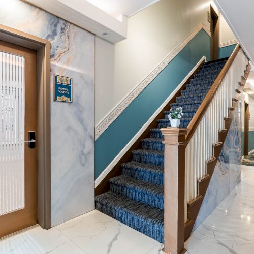 A hotel hallway with a staircase to the right, marble floors, blue carpet on the stairs, wall sconces, and a closed door on the left.
