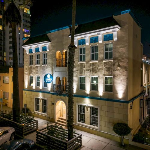 A stylish, cream-colored, three-story building at night with arched doorway, blue-lit windows, and palm trees in front.