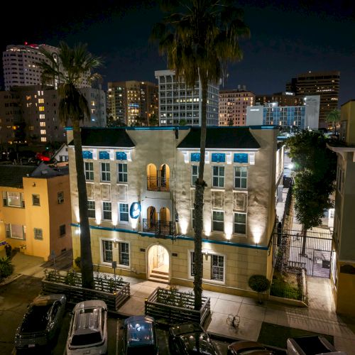 A night view of a small, well-lit building with a blue roof, palm trees, and surrounding colorful apartment blocks and parked cars, city lights glow.