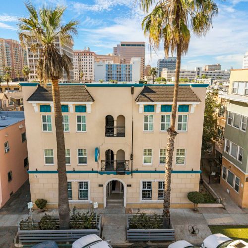 A pastel-colored, multi-story Mediterranean-style building with arched entry, palm trees, and nearby pastel townhouses under a blue sky.