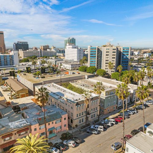 A sunny cityscape with mid-rise buildings, palm trees lining streets, and a mix of parked cars along a busy urban block.