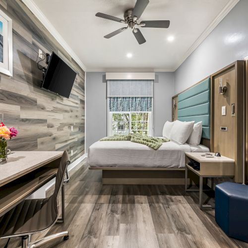 A modern hotel-style room with a comfy bed, wall-mounted TV, work desk, ceiling fan, large mirror, and a window with blinds; compact and bright.