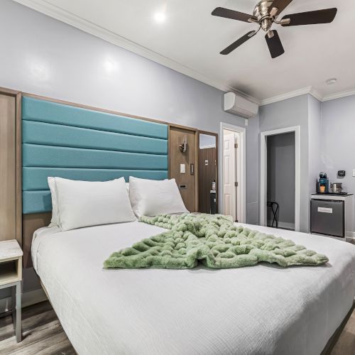 A modern hotel room with a large bed, teal upholstered headboard, two nightstands, ceiling fan, and a small desk area near the door. Always ending the sentence.