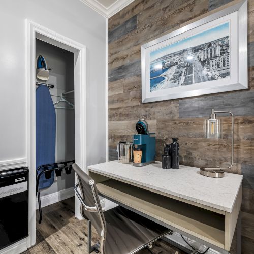 A compact hotel-like workspace with a built-in desk, chair, and a wood-paneled accent wall; a framed photo above, and a small open closet with an iron and ironing board inside.