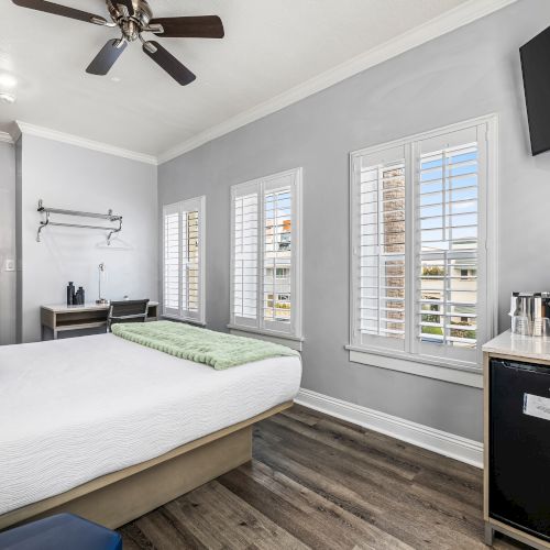 A bright hotel-style room with a bed, ceiling fan, multiple windows with white shutters, a small side table with a mini-fridge and coffee station, and a mounted TV.