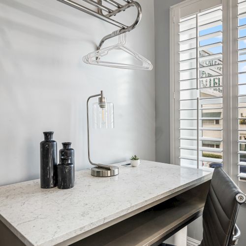 A tidy workspace with a marble countertop, hanging clothes rack, glass bottles, a white clipboard, and a chair by a window with blinds.