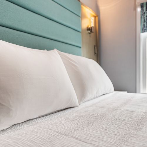 A tidy hotel bed with white sheets and two plump pillows, an upholstered teal headboard, soft lighting, and a bright window view nearby.