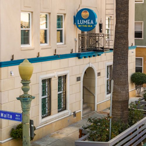 A sunny street scene of a Mediterranean-style hotel with white walls, blue trim, palm trees, and a sign for Lumea, Malta nearby.