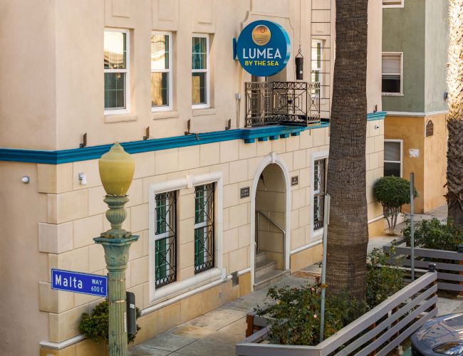 A sunny street scene of a Mediterranean-style hotel with white walls, blue trim, palm trees, and a sign for Lumea, Malta nearby.
