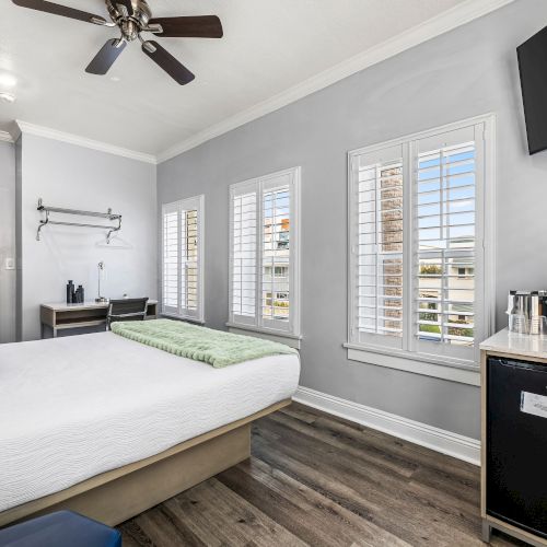 A modern hotel-style bedroom with a queen bed, ceiling fan, several windows with shutters, a wall-mounted TV, a small minibar, and a coffee station, all in gray tones.