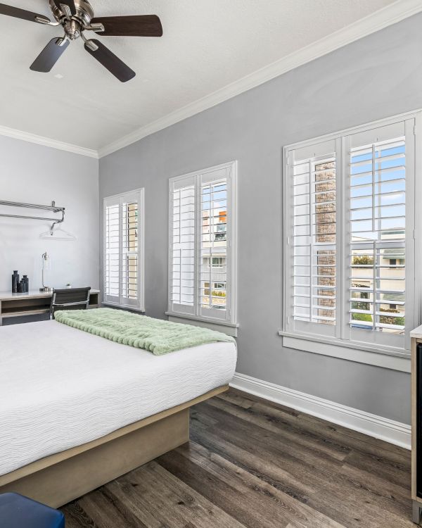 A modern hotel-style bedroom with a queen bed, ceiling fan, several windows with shutters, a wall-mounted TV, a small minibar, and a coffee station, all in gray tones.