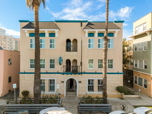 A peach-colored, three-story beach house with turquoise trim, arched entry, wrought-iron balconies, and tall palm trees in front.