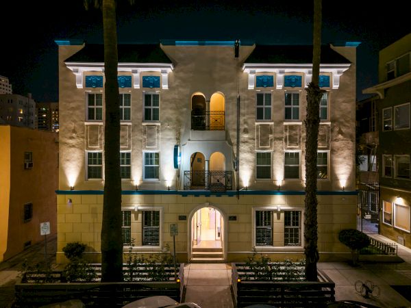 A Mediterranean-style building at night, illuminated facade with arched windows, central balcony, palm trees framing the front entrance, cozy urban setting.