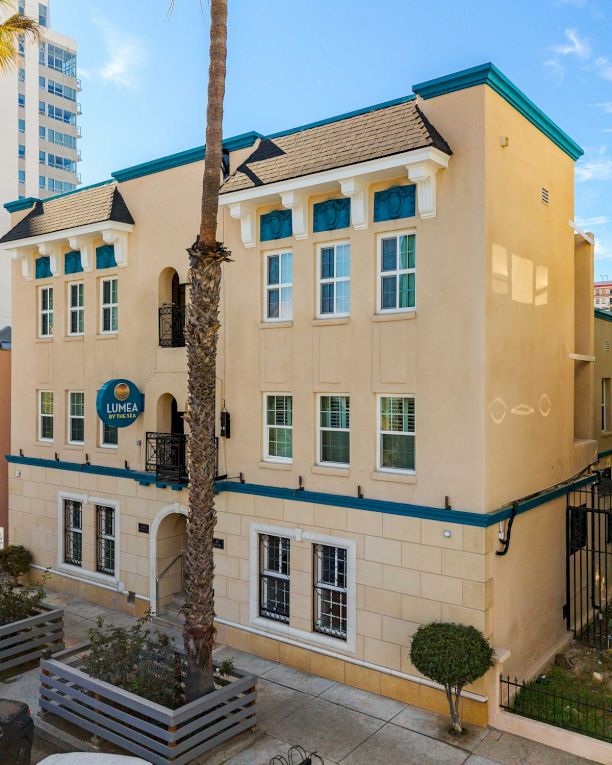 Colorful two-story Mediterranean-style building with cream walls, teal trim, palm trees, and a blue sky in a sunny urban setting.