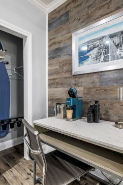 A small modern hotel-style workspace with a desk, chair, and framed artwork, a closet with an iron and luggage, and a wood-paneled accent wall.