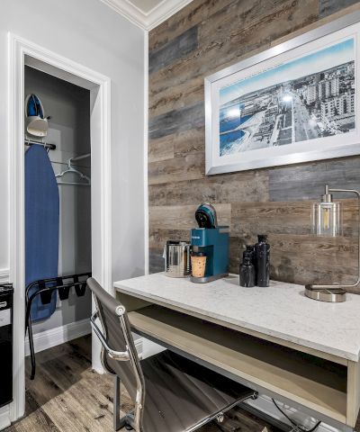 A small modern hotel-style workspace with a desk, chair, and framed artwork, a closet with an iron and luggage, and a wood-paneled accent wall.