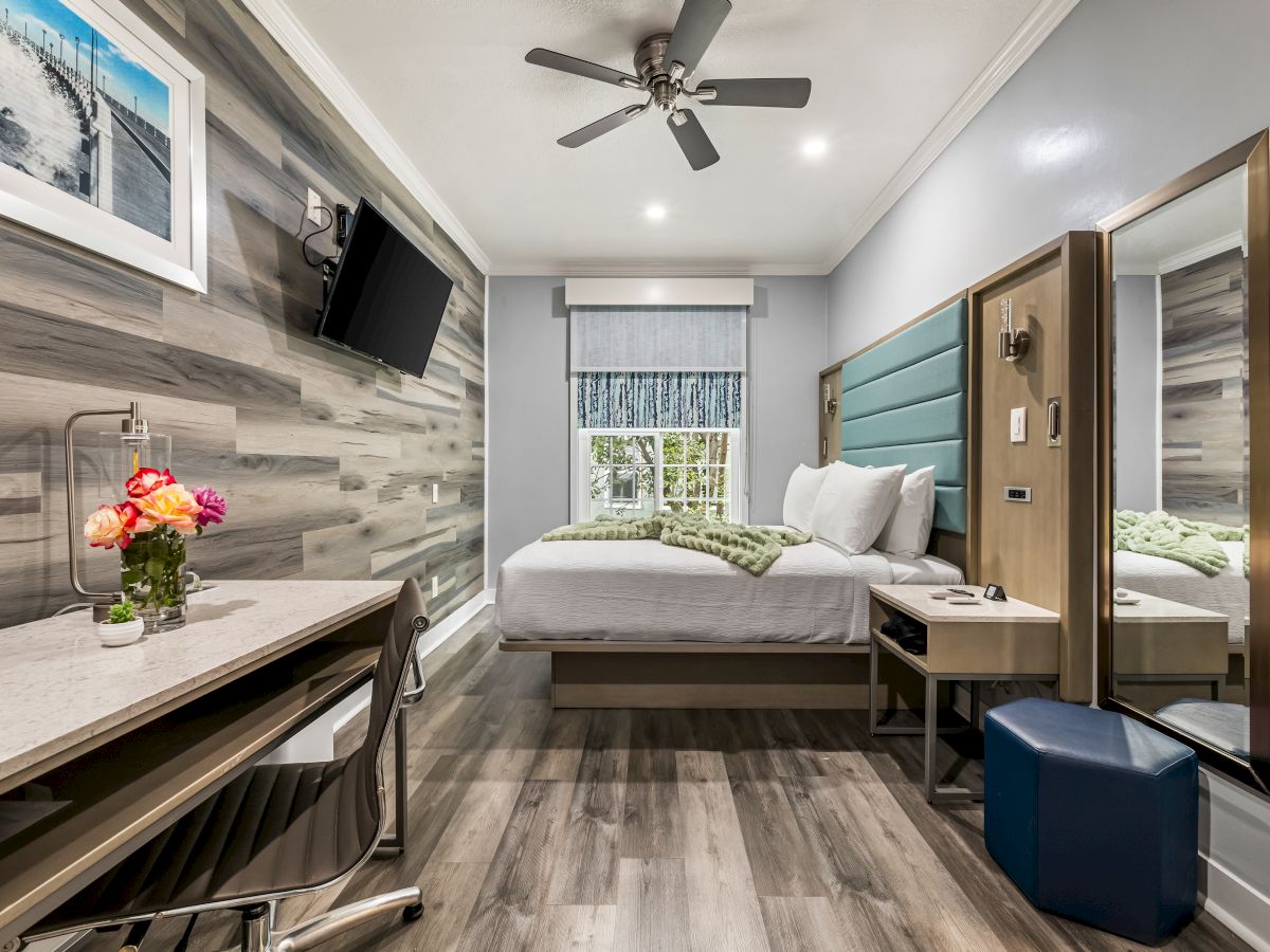 Modern hotel room with a king bed, wall-mounted TV, desk, ceiling fan, full-length mirror, and a blue upholstered headboard. Always ends the sentence.