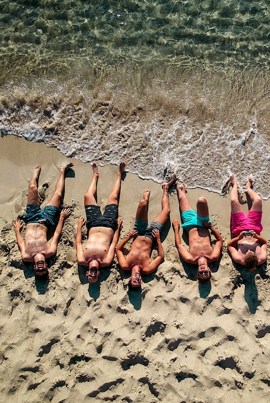 Six people lie on a sunny beach at the shoreline, facing upward, heads near the water, legs stretched out on the sand.