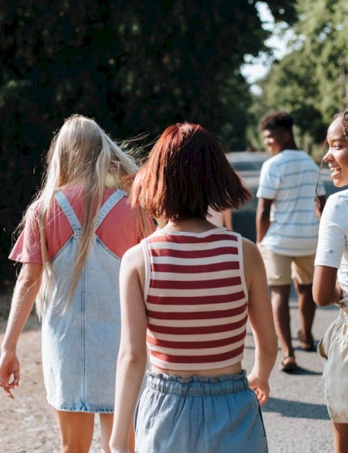A group of friends walking along a sunlit path, seen from behind, wearing casual summer clothes and chatting as they stroll.