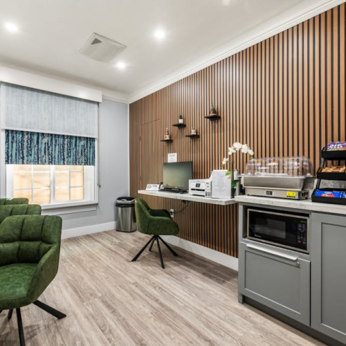 A modern, compact caf&eacute;-style workspace with a wooden slat wall, snack station, small desk, and green chairs in a bright, airy room.