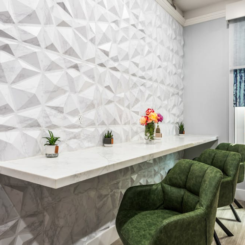 A modern interior with a geometric textured wall, a long marble counter, small potted plants, a vase of flowers, and green chairs by a window.