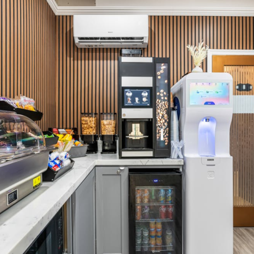 A compact, modern kitchenette with a coffee/tea station, a beverage fridge, a snack display, and a sleek water cooler beside a glass-front cabinet.
