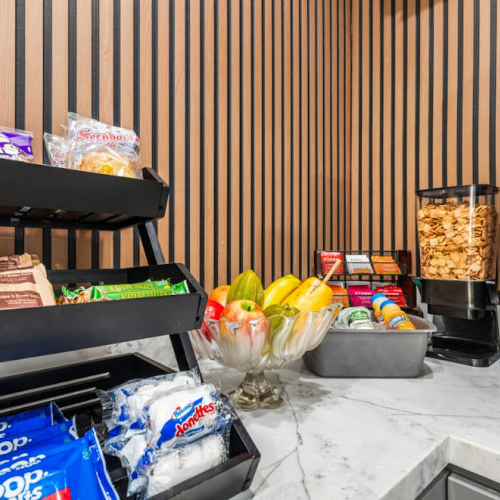 A snack bar with neatly organized shelves of chips, popcorn, and candy beside a bowl of fresh fruit on a marble counter.