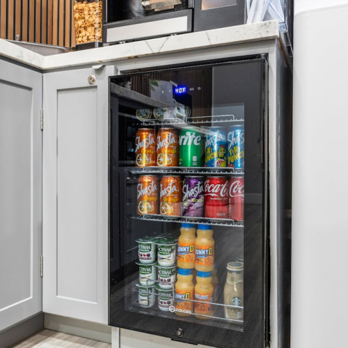A small built-in mini-fridge inside an open kitchen cabinet, stocked with assorted sodas and juice bottles on glass shelves.