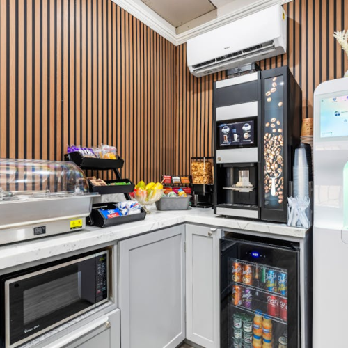 A compact break area with a vending fridge, beverage cooler, snack station, microwave, chiller, coffee/tea setup, and a white water dispenser in a corner, all against striped brown walls.