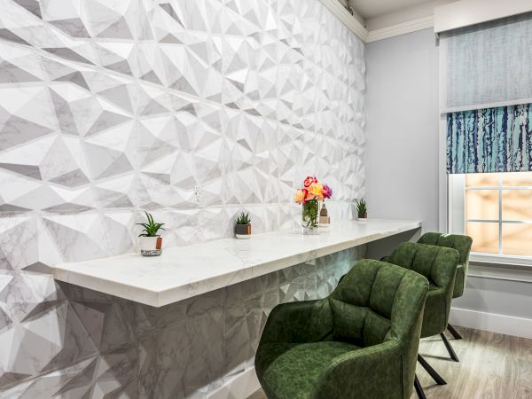 A modern interior with a textured white geometric wall, a long white counter, green upholstered chairs, small plants, and a window with a patterned blind.